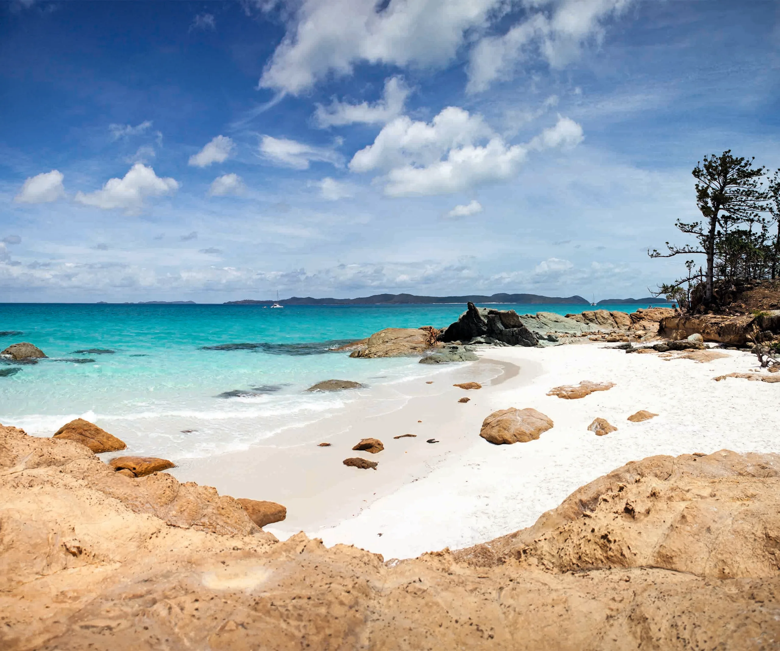 Rocky beach with turquoise ocean, clear blue sky, and distant hills under scattered clouds.