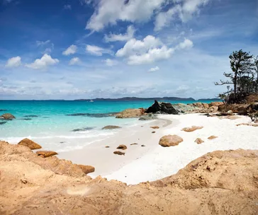 Rocky beach with turquoise ocean, clear blue sky, and distant hills under scattered clouds.