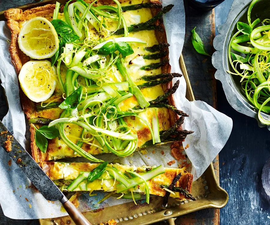Asparagus and lemon tart garnished with fresh herbs, served on a tray, with a side bowl of greens.
