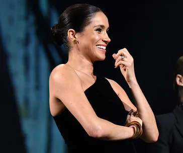 A woman in a black dress smiles and gestures on stage, wearing gold earrings and bracelets.
