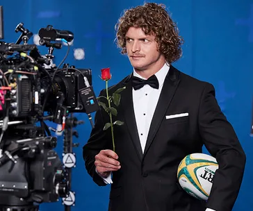 Man in tuxedo holds a rose and rugby ball, standing by a camera against a blue background.