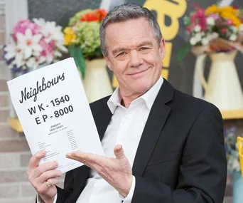 Paul Robinson holds the 8000th episode script of "Neighbours," surrounded by flowers and smiling confidently.