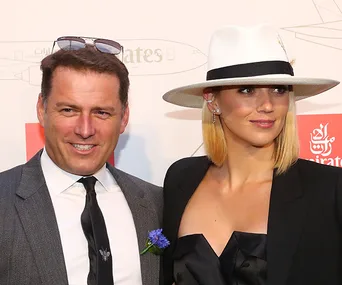 Two people in formal attire at an event; man in suit, woman in hat and black dress, standing together and smiling.