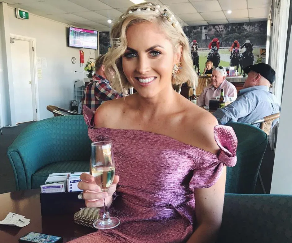 Woman in a pink off-shoulder dress, smiling and holding a champagne glass, seated in a social setting.