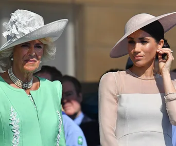 Camilla and Meghan Markle at an outdoor event, both wearing elegant hats and formal dresses.