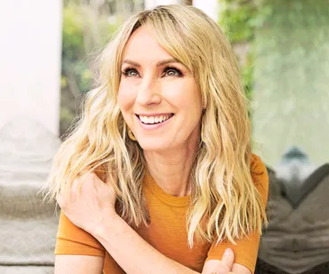 A smiling person with long blonde hair wearing an orange shirt, sitting in a bright, softly lit space.
