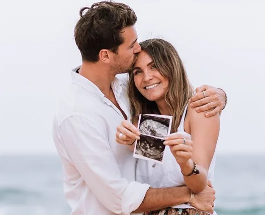 Couple embracing on a beach, woman holding an ultrasound photo, both smiling and looking joyful.