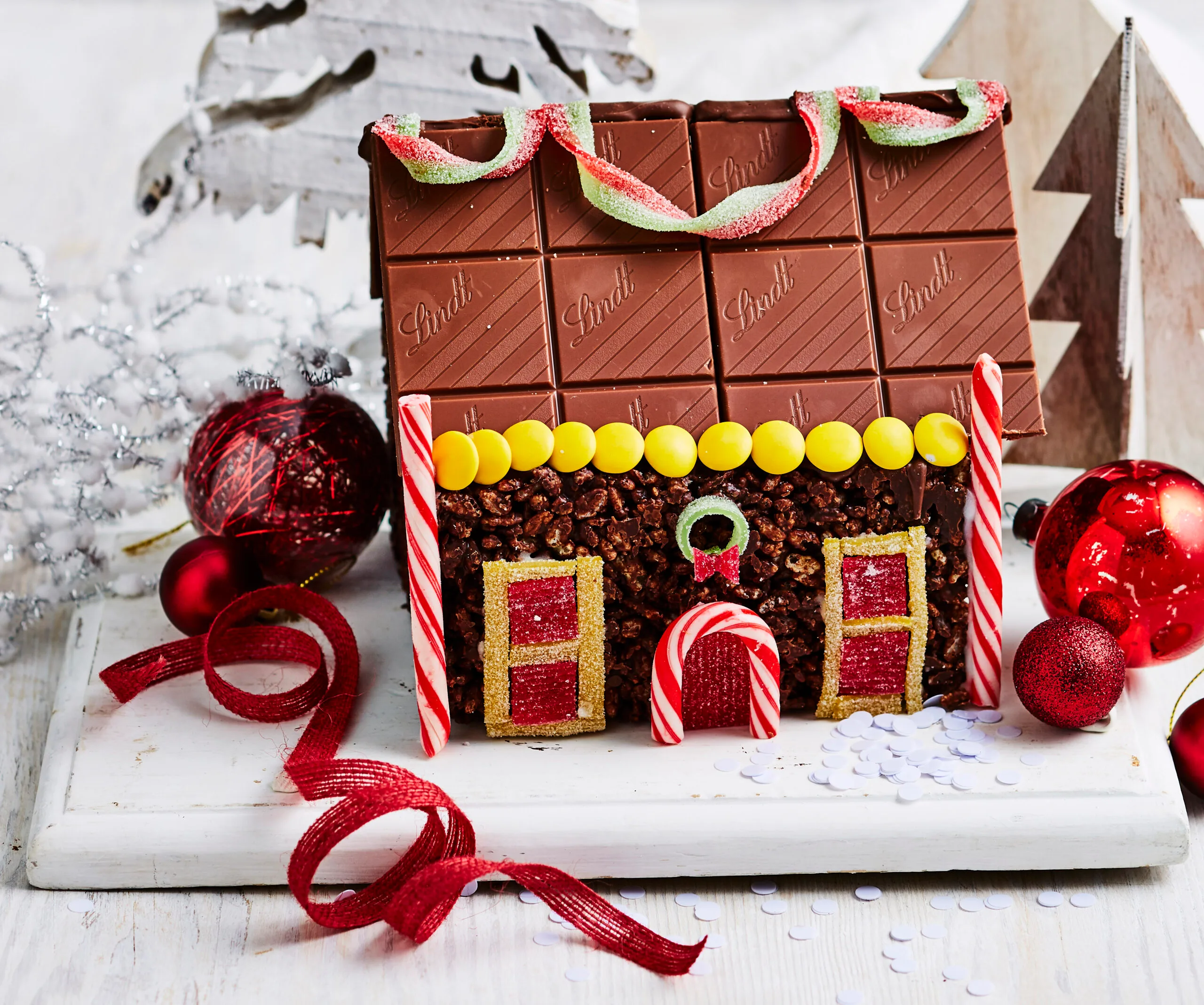 Chocolate gingerbread house with candy decorations, surrounded by Christmas ornaments and ribbons.