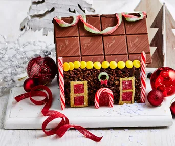 Chocolate gingerbread house with candy decorations, surrounded by Christmas ornaments and ribbons.