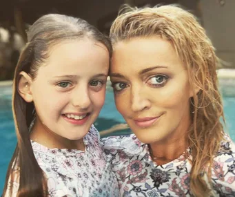 A woman and a young girl smiling together in front of a pool, both wearing floral-patterned outfits.
