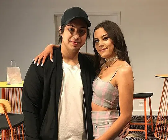 Two people posing side by side indoors, one in a cap and jacket, the other in a plaid outfit, smiling at the camera.