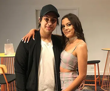 Two people posing side by side indoors, one in a cap and jacket, the other in a plaid outfit, smiling at the camera.