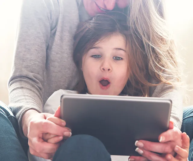 A child with surprised expression watches tablet while sitting with an adult.