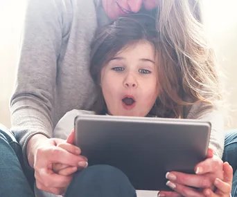 A child with surprised expression watches tablet while sitting with an adult.