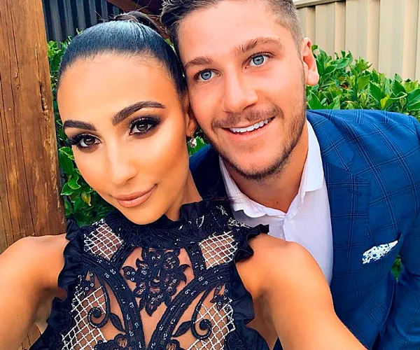 A couple taking a selfie outdoors, with the woman in a stylish black lace outfit and the man in a blue suit.