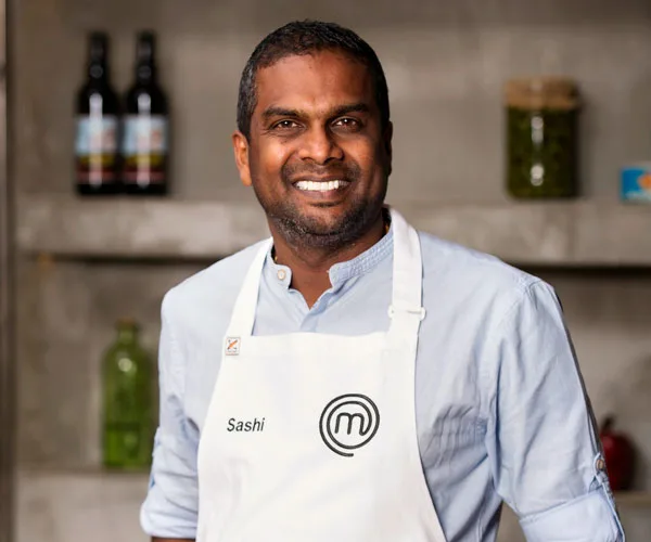 Sashi Cheliah smiling in a kitchen, wearing a MasterChef apron.