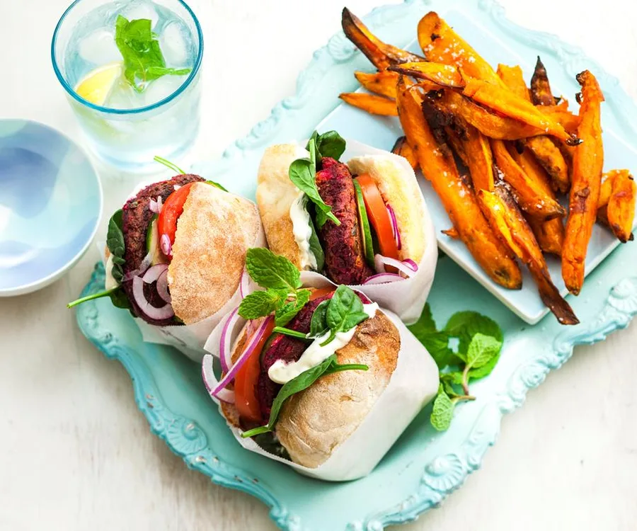 Vegan burgers wrapped in paper on a tray with sweet potato fries and a glass of lemon water, garnished with mint.