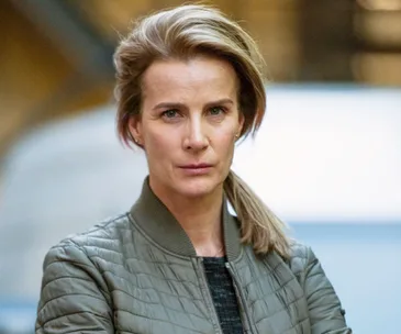 Rachel Griffiths in a green jacket, serious expression, standing outdoors with blurred background.