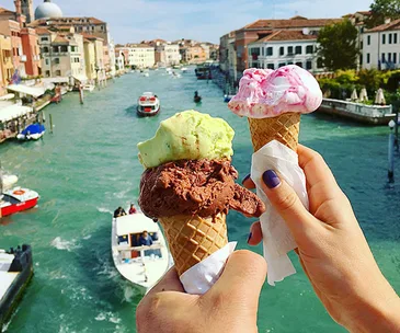 Two hands holding ice cream cones over a canal in Venice, Italy, with boats and historic buildings in the background.