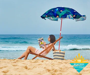 Woman relaxing under an umbrella on beach, reading magazine, with ocean waves in the background. "19 Beaches" logo visible.