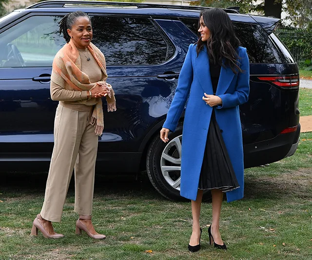 Two women standing outside near a parked blue SUV, one in a beige outfit with a scarf, the other in a blue coat.
