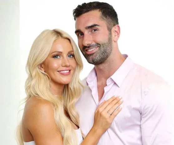 A smiling couple posing together, the woman with blonde hair and the man in a light shirt, both seeming happy.