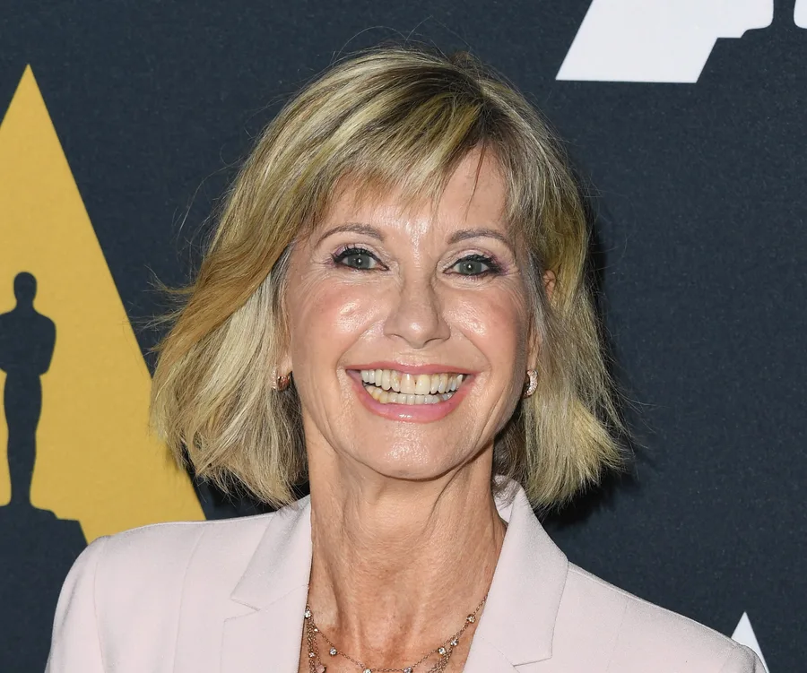 "Smiling woman with short blonde hair in a light-colored blazer against a dark backdrop."