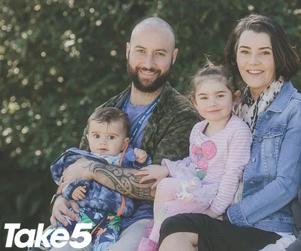 A family of four sitting outdoors, smiling; father holding baby, mother and young girl beside him. "Take 5" logo in corner.