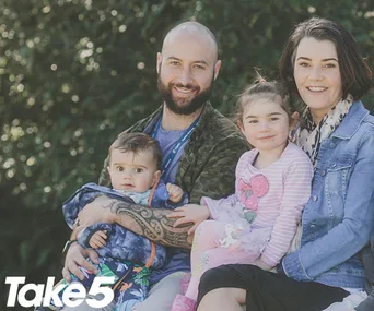 A family of four sitting outdoors, smiling; father holding baby, mother and young girl beside him. "Take 5" logo in corner.