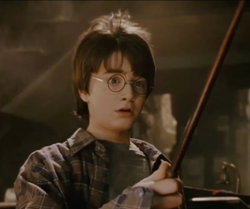 Young boy holding a magic wand appears surprised, wearing round glasses and a plaid shirt, in a dimly lit room.