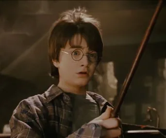 Young boy holding a magic wand appears surprised, wearing round glasses and a plaid shirt, in a dimly lit room.