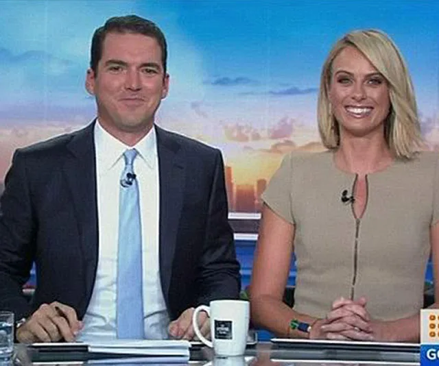 Two news anchors sitting at a desk, smiling, with microphones clipped to their clothing.
