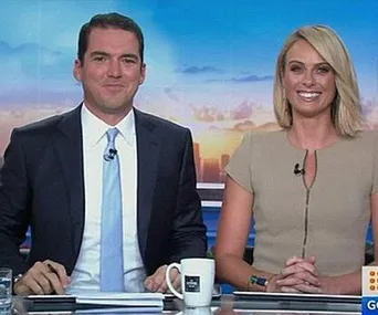 Two news anchors sitting at a desk, smiling, with microphones clipped to their clothing.