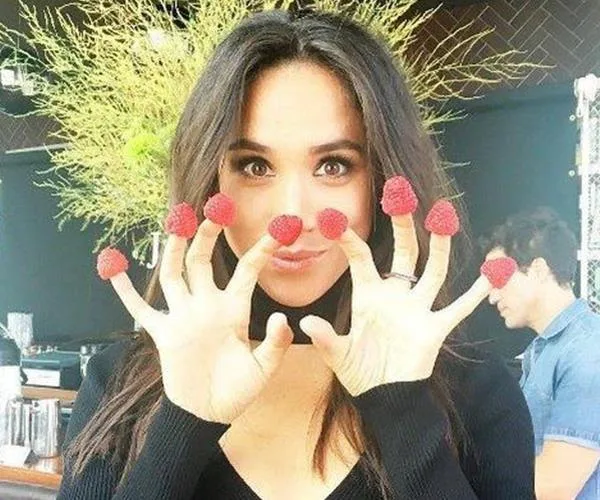 Woman playfully poses with raspberries on fingertips, smiling.