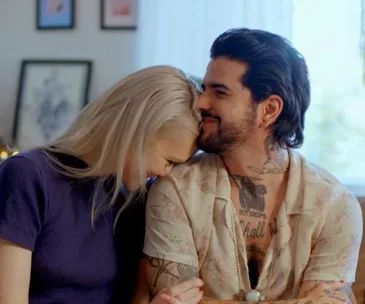 A couple sits closely; the woman with blonde hair leans on the tattooed man, both smiling warmly.