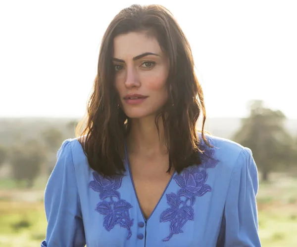 A woman with wavy hair in a blue embroidered blouse stands outdoors, softly lit by natural light, with trees in the blurred background.