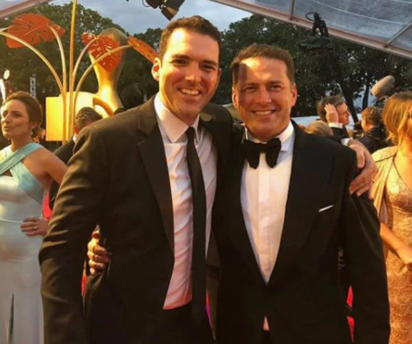 Two men in suits pose together at a formal event with people in evening attire in the background.