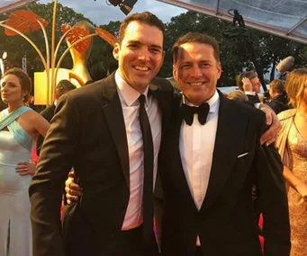 Two men in suits pose together at a formal event with people in evening attire in the background.