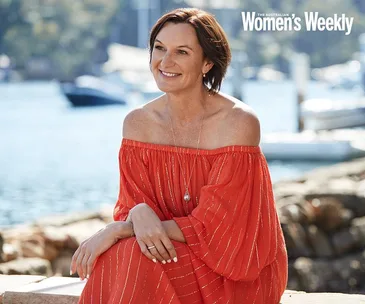 Woman in a red dress sits by the waterfront, smiling, with blurred boats and trees in the background.