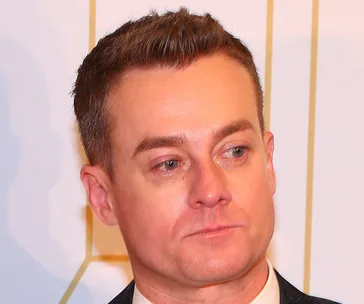 Man with short brown hair in a suit at an event, looking to the side with a neutral expression.