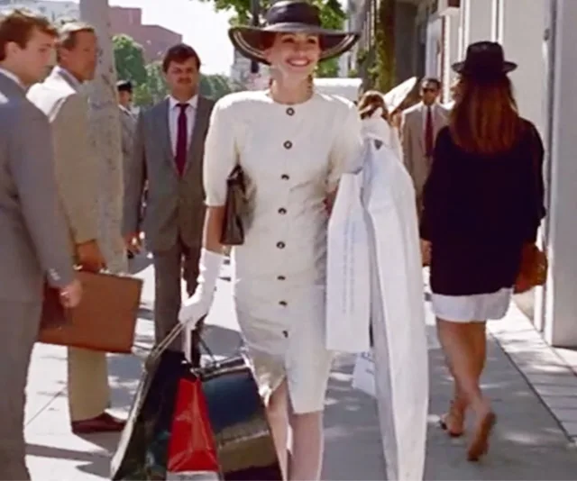 "Person in a stylish white dress and hat, holding shopping bags while smiling and walking on a busy street."