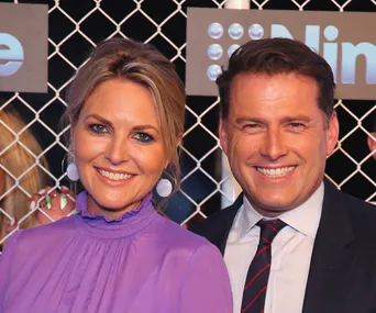 "Two people smiling in front of a chain-link backdrop with 'Nine' branding, one wearing a purple dress, the other in a suit."
