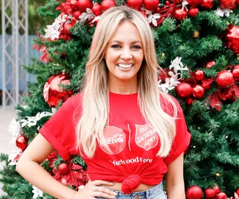 Person in a red Christmas-themed shirt smiling in front of a decorated tree with red ornaments.