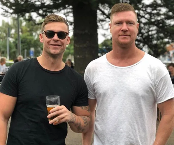 Two men standing outside, one in a black shirt holding a drink, the other in a white shirt. Both appear relaxed and smiling.