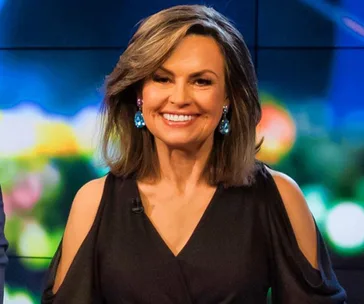 Lisa Wilkinson smiling on a television set, wearing a black dress with cut-out shoulders and turquoise earrings.