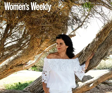 A woman in a white lace top stands by a tree in a sunny outdoor setting, looking away.
