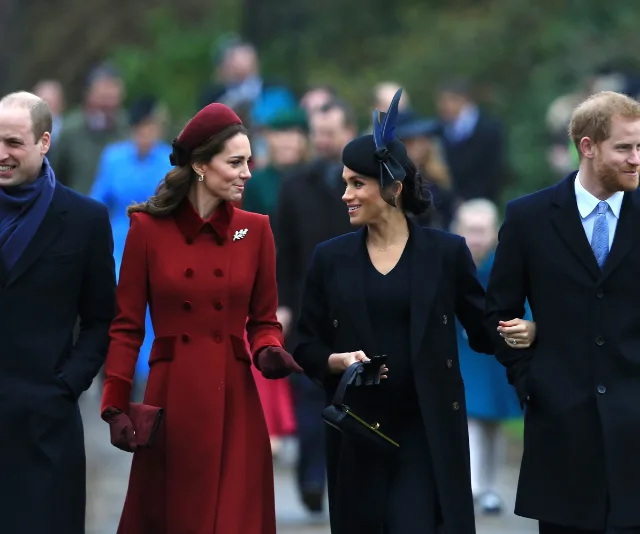 Two women in coats, one red and one black, smile at each other while arm-in-arm with two men in suits outdoors.