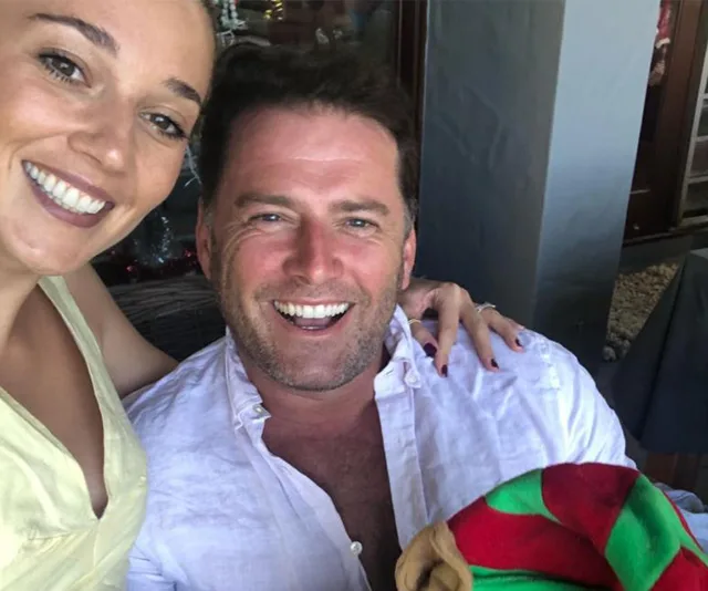 Man and woman smiling for a selfie, man wearing a white shirt, woman in yellow, indoor setting.