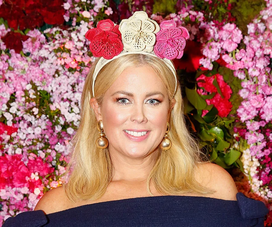 A woman with blonde hair wearing a floral headband and pearl earrings, in front of a backdrop of pink flowers.