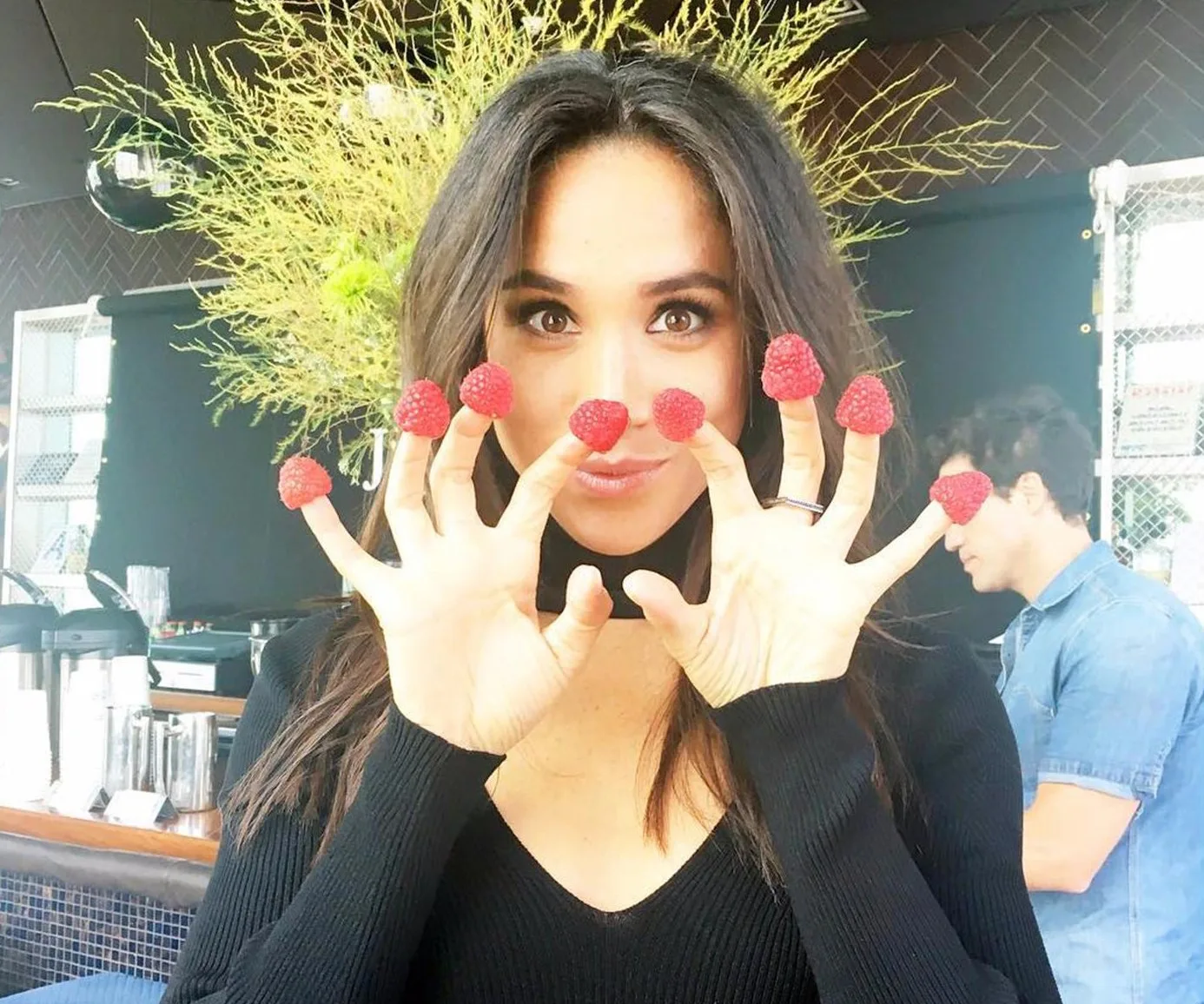 A woman playfully holds raspberries on her fingers, smiling in a casual setting.